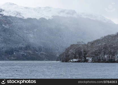 Beautiful Winter landscape image of snow covered trees on shores of Loch Lomond with Ben Lomond mountain looming in background