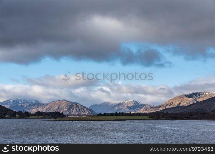 Beautiful Winter landscape view along Loch Leven past Ballachulish Bridge towards snowcapped mountains in distance with dramatic sky