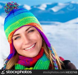 Beautiful winter portrait of woman, wearing cute colorful hat and scarf standing on snowy mountains background, wintertime holidays