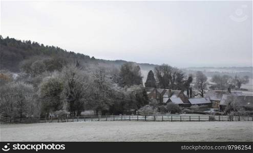 Beautiful Wintry landscape of farm in fog in English countryside at dawn with hoarfrost covering trees and buildings