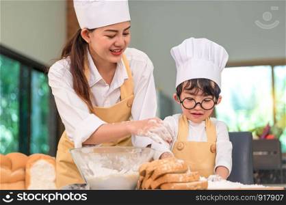 Beautiful woman and cute little Asian boy with eyeglasses, chef hat and apron playing and baking bakery in home kitchen funny. Homemade food and bread. Education and learning concept. Thai person