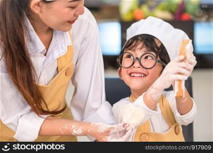Beautiful woman and cute little Asian boy with eyeglasses, chef hat and apron playing and baking bakery in home kitchen funny. Homemade food and bread. Education and learning concept. Thai person