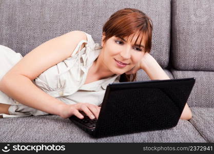 Beautiful woman at home lying on sofa and working with a laptop