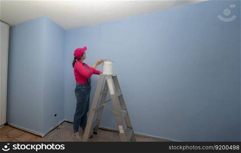 Beautiful woman dressed in overalls and red t-shirt going up a metal ladder with a jar of paint next to a blue wall in an empty room. woman dressed in overalls and red t-shirt going up a metal ladder with a jar of paint next to a blue wall in an empty room