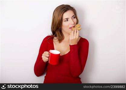 Beautiful woman drinking a hot coffee and eating a gingerbread cookie, against a gray wall