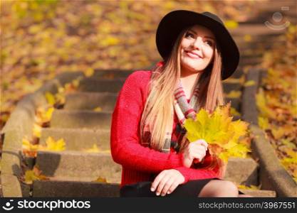 Beautiful woman in autumn park. Portrait of attractive young smiling woman holding big gold leafs in autumnal park. Fashionable girl wearing red sweater scarf and black hat.