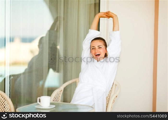 Beautiful woman in bathrobe sitting at table on terrace and stretching after sleeping&#xA;