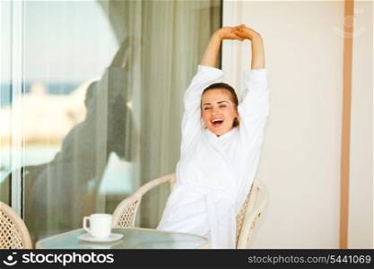 Beautiful woman in bathrobe sitting at table on terrace and stretching after sleeping&#xA;
