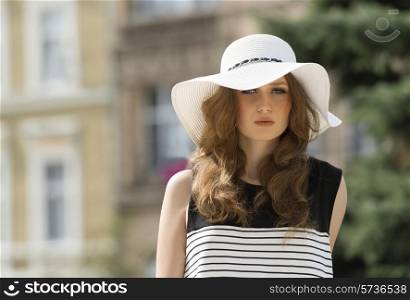 Beautiful, woman in beautiful striped dress and white summer hat on the street in summer day. She has got brown, long, curly hair.