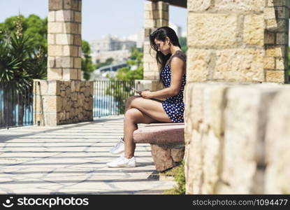 Beautiful woman in blue dress sitting on a bench while using a smartphone