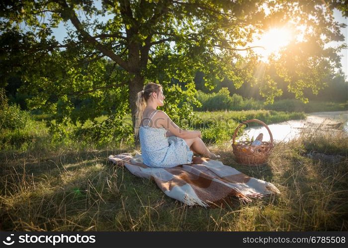 Beautiful woman in dress relaxing on blanket under tree and looking at sunset