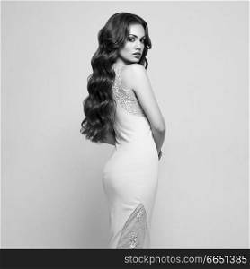 Beautiful Woman in Elegant white Evening Dress. Brunette Girl with Long Healthy and Shiny Curly Hair. Care and Beauty. Beautiful Model Woman with Wavy Hairstyle