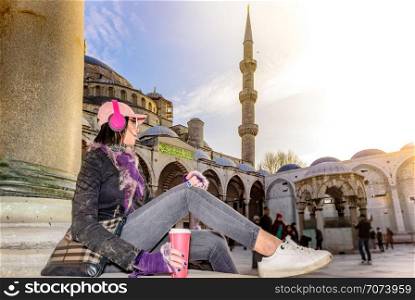 Beautiful woman in hat with takeaway coffee sunglasses,headphones sits at courtyard of Sultanahmet Mosque or Blue Mosque in Istanbul,Turkey. Beautiful woman traveler stands in front of Bosphorus,a popular destination in Istanbul,Turkey