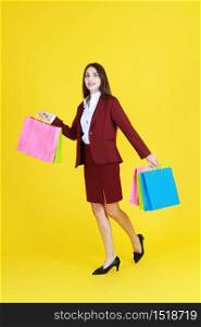 Beautiful woman in red suit smile and hand holding colorful shopping paper bags isolated on yellow background.