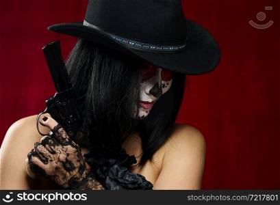 beautiful woman in skeleton make-up holds a revolver, she is wearing a black hat, red background