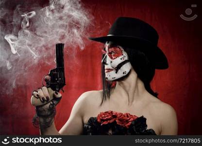 beautiful woman in skeleton make-up holds a revolver, she is wearing a black hat. White smoke over red background from shot