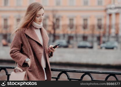 Beautiful woman in warm coat, holds modern cell phone, messages in social networks, connected to free wifi, walk in city, street style, blurred background with copy space for your advertisement