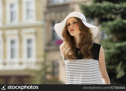 Beautiful woman in white summer hat. She has curly, long, brown hair. Her makeup is so soft. She wears striped dress.