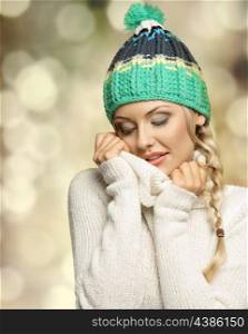beautiful woman in winter dress with green hat in act to warm sweetly herself with white wool sweater . keeping closed eyes