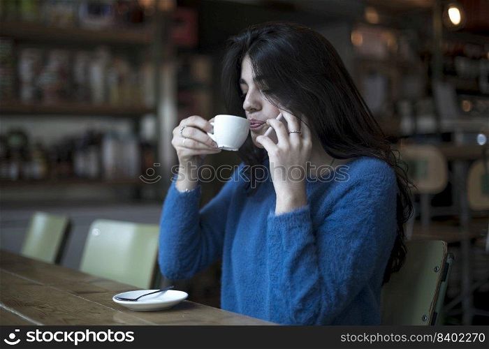 Beautiful woman is drinking delicious coffee while sitting in the bar while talking by phone