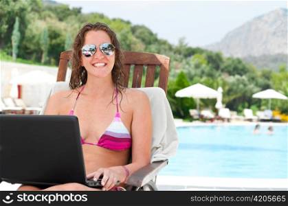 beautiful woman is sitting on wooden chair near the pool with laptop