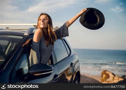 Beautiful woman looking out the car window waving with a hat, near the beach