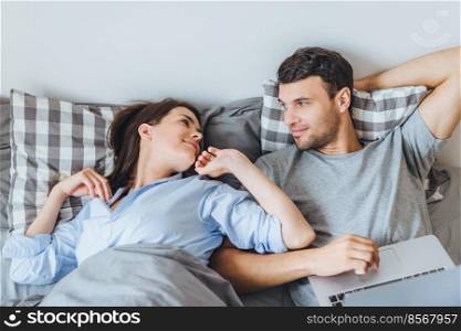 Beautiful woman looks with great love at her husband, just awake, says good morning and male who keyboards on laptop computer, happy to see each other. Leisure and relationship concept