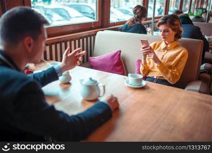 Beautiful woman makes selfie on phone camera against man in suit, restaurant on background. Man and woman happy together. Woman makes selfie on camera against man