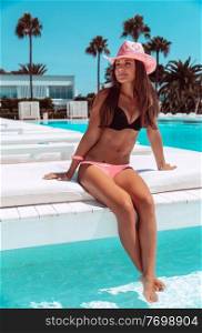 Beautiful woman on the beach resort, attractive model relaxing on luxury white sunbed and gladly wets her feet in the cool water of the pool, enjoying summer vacation, Cyprus