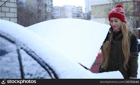 Beautiful woman opening trunk of her car, covered with snow, and taking snow brush to clean her automobile on winter morning.