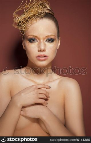 beautiful woman posing in beauty portrait with creative golden make-up, accessory in the hair-style. She is covering her naked breast and looking in camera &#xA;