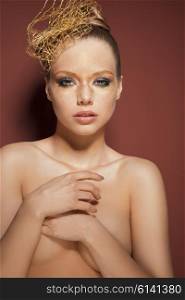 beautiful woman posing in beauty portrait with creative golden make-up, accessory in the hair-style. She is covering her naked breast and looking in camera &#xA;
