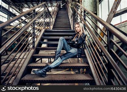 Beautiful woman posing wearing casual outfit with leather jacket, black shoes and fashionable jeans. Girl posing in industrial plant.