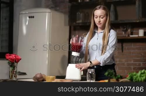 Beautiful woman pouring vegetable smoothie from blender shaker jug into glass mason jar in the kitchen. Attractive female making healthy organic food and pouring beet smoothie from blender to glass.