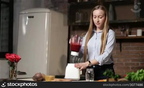 Beautiful woman pouring vegetable smoothie from blender shaker jug into glass mason jar in the kitchen. Attractive female making healthy organic food and pouring beet smoothie from blender to glass.