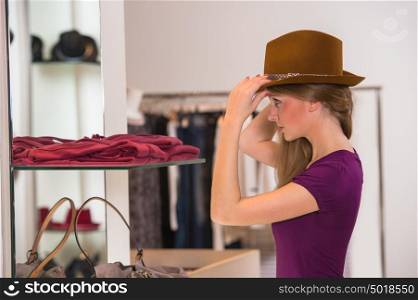 Beautiful woman shopping in clothing store trying on brown hat in front of mirror
