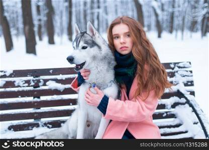 Beautiful woman sitting on the bench with siberian husky, snowy forest on background. Cute girl hugs with charming dog. Real friendship with domestic animal. Woman sitting on the bench with siberian husky
