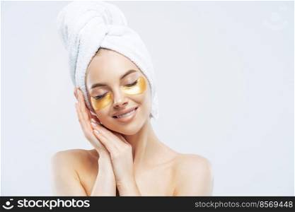 Beautiful woman stands with closed eyes, has soft healthy skin, leans at hands near face, smiles gently, wears golden patches to reduce puffiness under eyes, stands shirtless, wears towel, takes bath