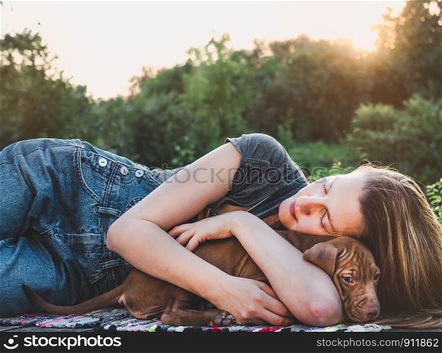 Beautiful woman tenderly hugging a young, charming puppy. Relax on the terrace against a sunset and green trees. Close-up. Concept of care, education, obedience training and raising of pets. Beautiful woman tenderly hugging a young puppy