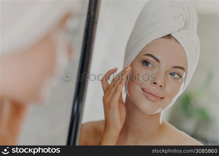 Beautiful woman touches face gently, looks at her reflection in mirror, enjoys softness of skin, stands with wrapped bath towel on head, has minimal makeup and manicure. Beauty treatment concept