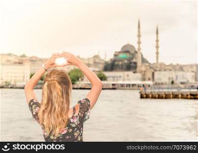Beautiful woman traveler makes heart shape with hands on view of New Mosque and Galata Bridge,popular destinations in Istanbul,Turkey. Beautiful woman traveler makes heart shape over New Mosque