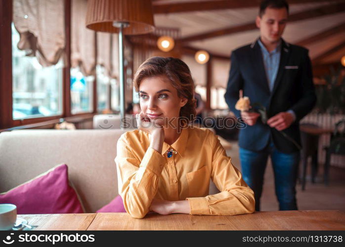 Beautiful woman waiting in restaurant, man with rose in hand on background