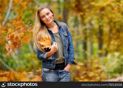 Beautiful woman walking in autumn park with dry tree leaf in hand, enjoying fall vacation, day off in the forest
