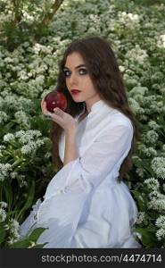 Beautiful woman wearing a long white dress sitting in white flowers holding a red apple