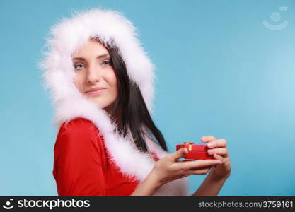 Beautiful woman wearing santa claus costume clothes holding small red gift box with gold bow on blue background. Christmas time gifts.