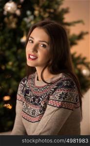Beautiful woman wearing winter outfit sitting on couch at home near Christmas tree