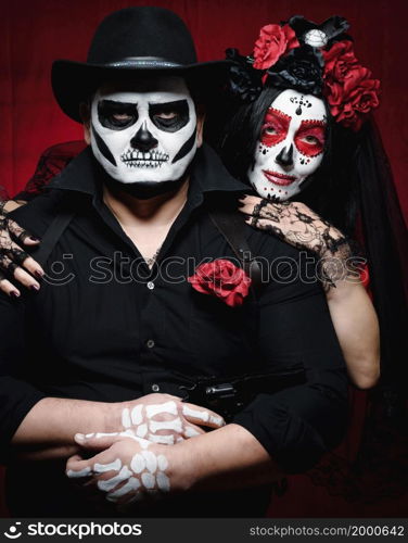 beautiful woman with a sugar skull makeup with a wreath of flowers on her head and a skeleton man in a black hat. Couple on dark red background