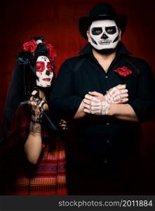 beautiful woman with a sugar skull makeup with a wreath of flowers on her head and a skeleton man in a black hat holding a gun