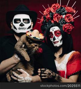 beautiful woman with a sugar skull makeup with a wreath of flowers on her head and a skeleton man in a black hat holding a gray cat