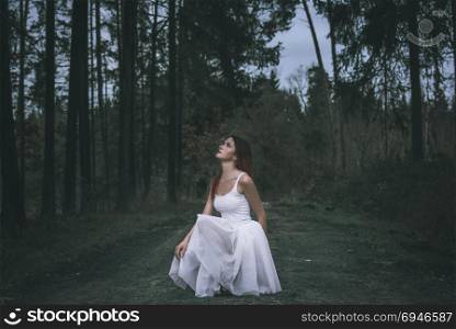 Beautiful woman with an ombre hairstyle, dressed in a white tutu skirt, in a squat position, on a forest road, looking up, in a spring woodland.